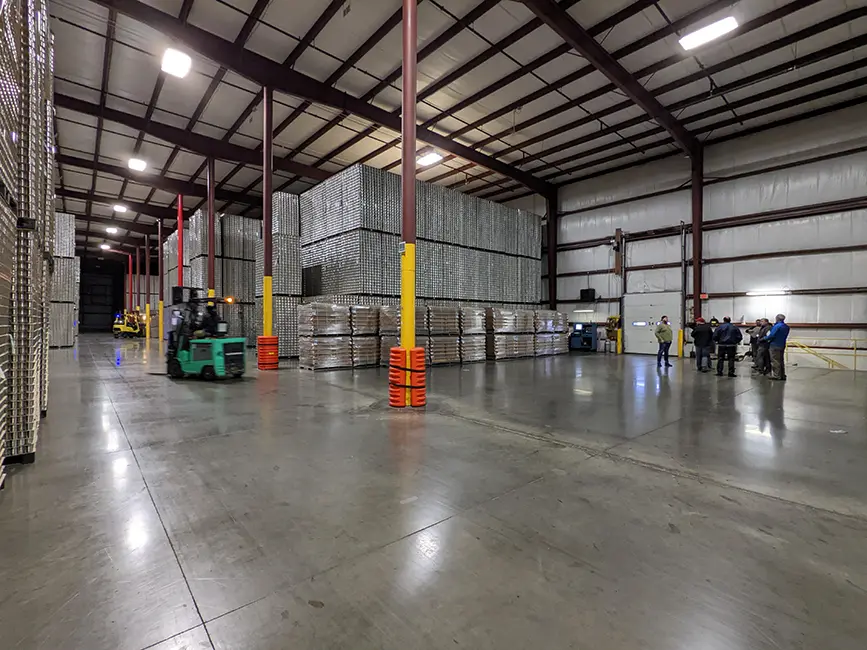 Wide view of an industrial warehouse interior with stacked aluminum cans, forklifts, and a group of workers.